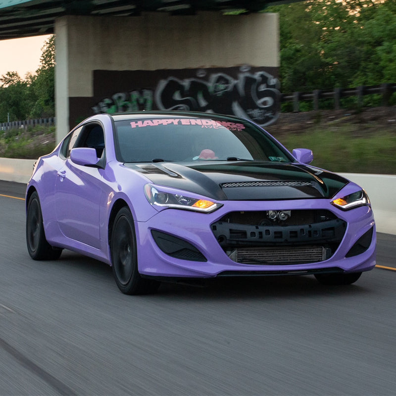 A rolling shot of a modified Hyundai Genesis Coupe featuring a gloss pearl lavender vinyl wrap and a vented carbon fiber hood.