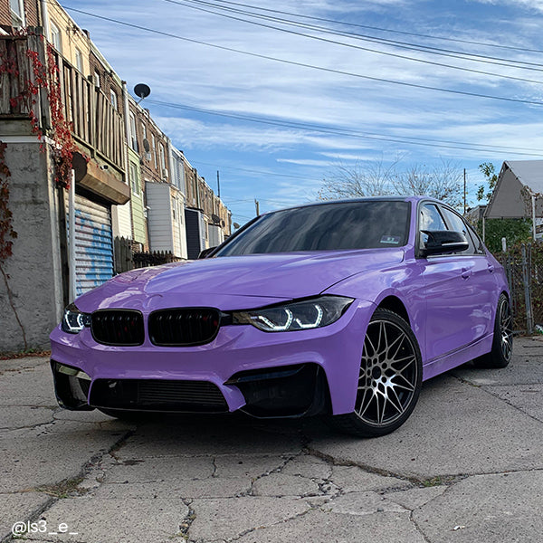 Front three-quarter view of a modified BMW 3-Series showcasing a full gloss pearl lavender vinyl wrap and custom black wheels in a city alley.