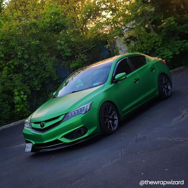 A high-angle shot of a lowered Acura ILX with a Satin Cheetah Green Metallic Vinyl Car Wrap and a front lip spoiler, set against a backdrop of lush trees.