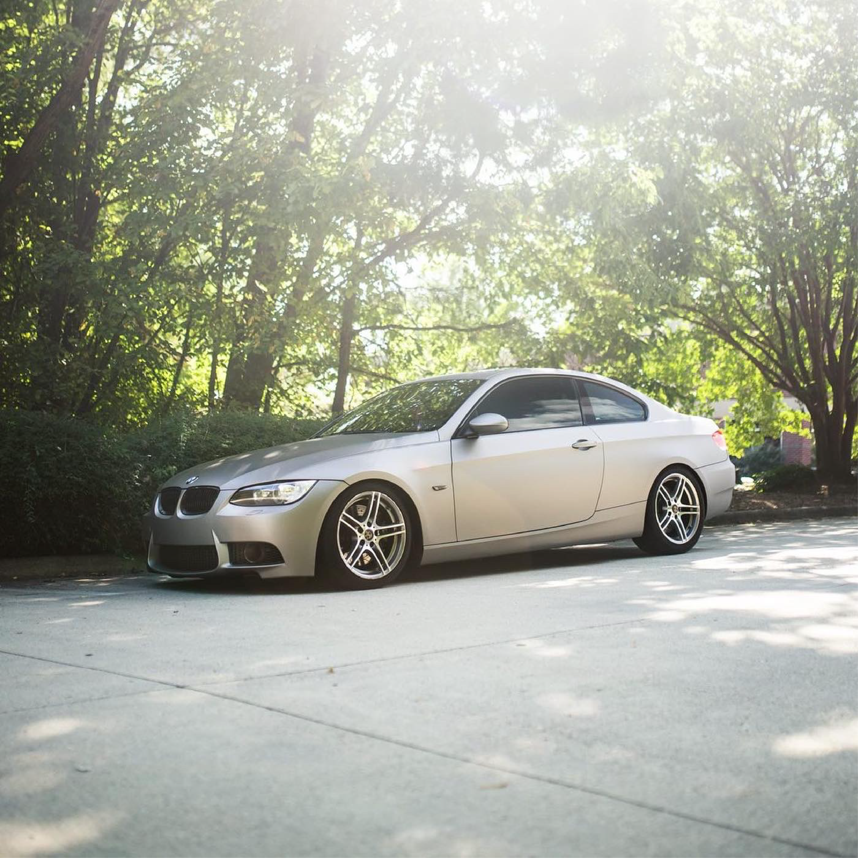 BMW wrapped in satin gray car wrap with black wheels, parked on asphalt in front of tall evergreen trees.