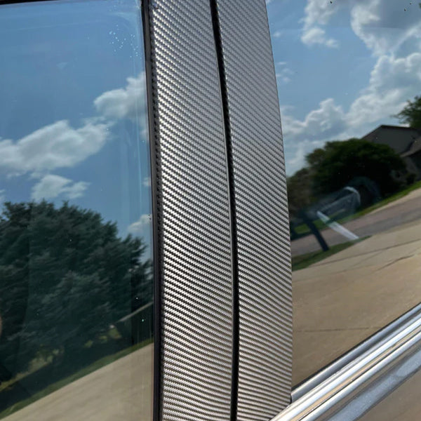 Close-up of a carbon fiber vinyl wrap on a vehicle's B-pillar, reflecting a clear sky and neighborhood backdrop.