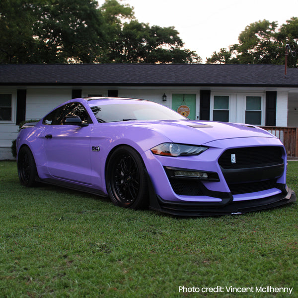 A widebody Ford Mustang with a full gloss pearl lavender vinyl wrap and a large front splitter, parked on a suburban lawn.