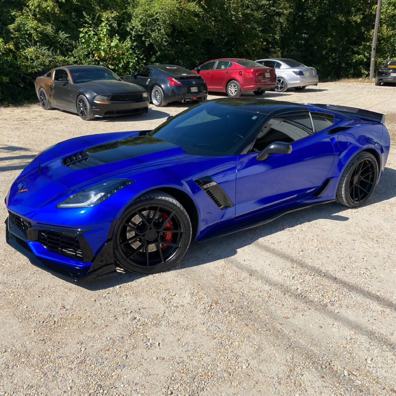Blue sports car parked on a gravel surface with other cars in the background.