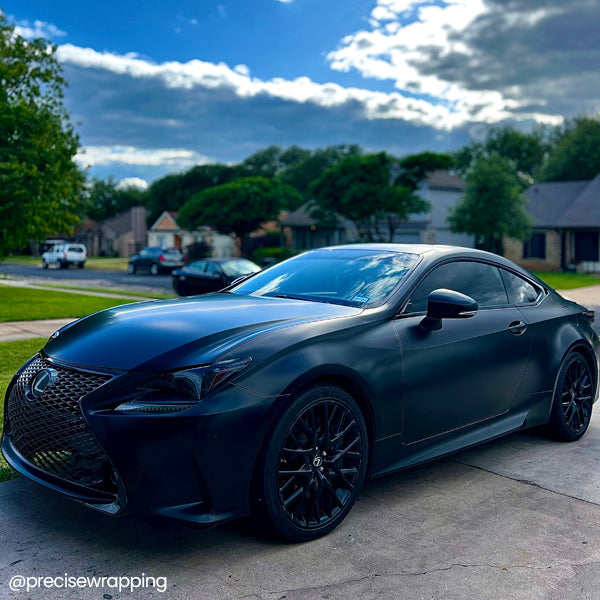 Lexus RC coupe wrapped in satin black car wrap with gloss black wheels, parked in a suburban neighborhood under a vibrant sky.