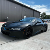 BMW i8 sports car wrapped in satin black car wrap with blue trim accents and black wheels, parked outside an industrial black metal building.