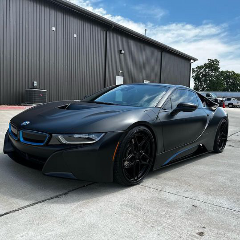 BMW i8 sports car wrapped in satin black car wrap with blue trim accents and black wheels, parked outside an industrial black metal building.