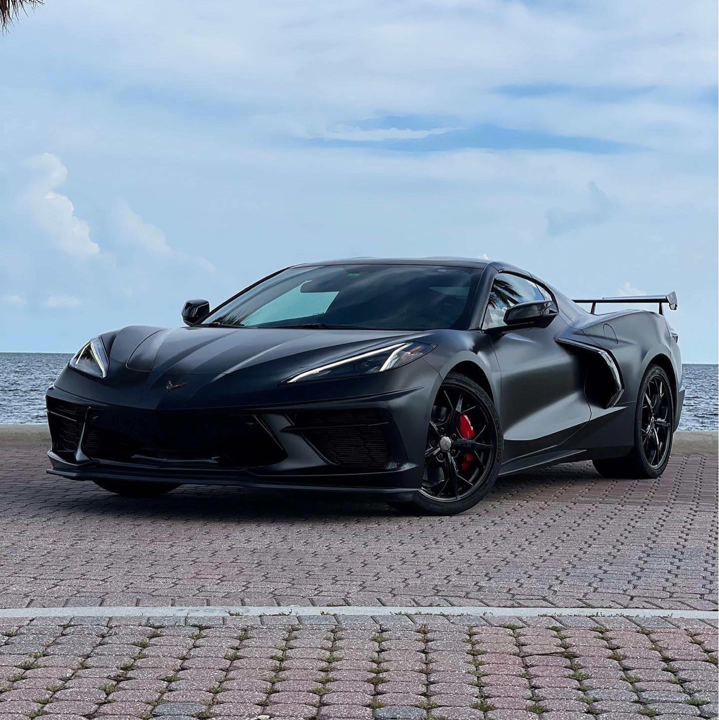 Chevrolet Corvette wrapped in satin black car wrap with red brake calipers, parked on a waterfront brick lot under a blue sky.
