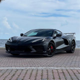Chevrolet Corvette wrapped in satin black car wrap with red brake calipers, parked on a waterfront brick lot under a blue sky.