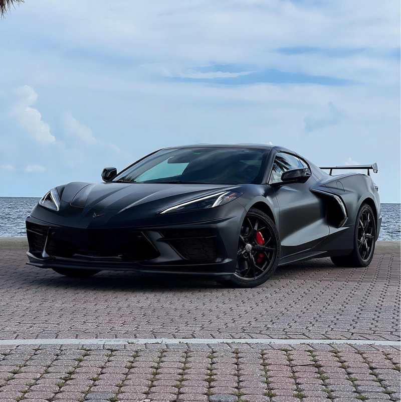 Chevrolet Corvette wrapped in satin black car wrap with red brake calipers, parked on a waterfront brick lot under a blue sky.