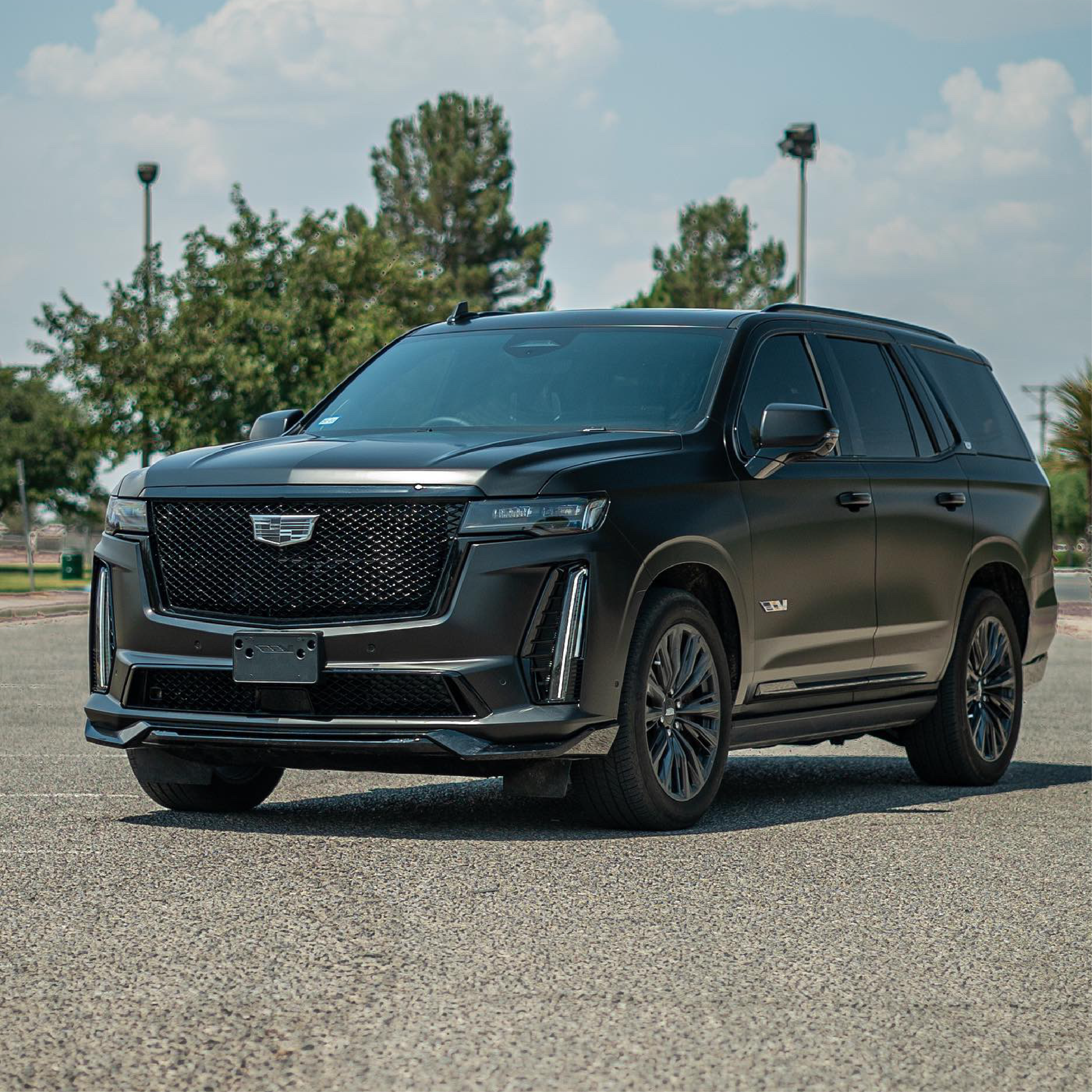Cadillac Escalade V-Series SUV wrapped in satin black car wrap, parked in a sunny open lot with trees and lamp posts in the background.