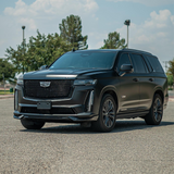 Cadillac Escalade V-Series SUV wrapped in satin black car wrap, parked in a sunny open lot with trees and lamp posts in the background.