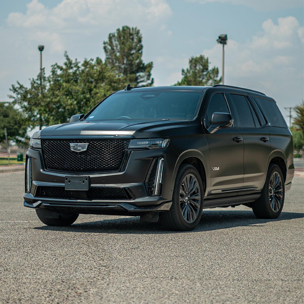 Cadillac Escalade V-Series SUV wrapped in satin black car wrap, parked in a sunny open lot with trees and lamp posts in the background.