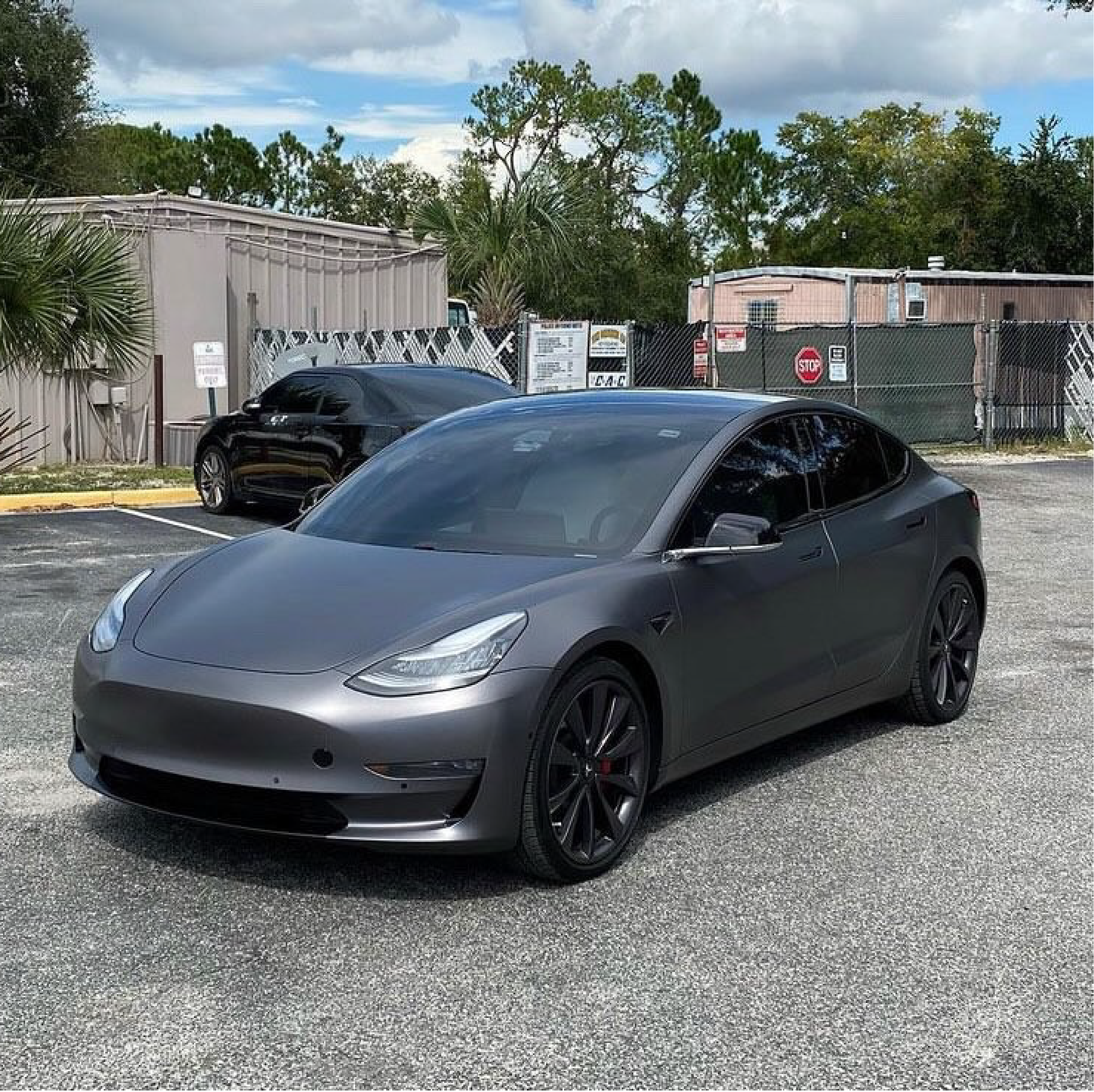 Gray Tesla car parked in a lot with trees and buildings in the background