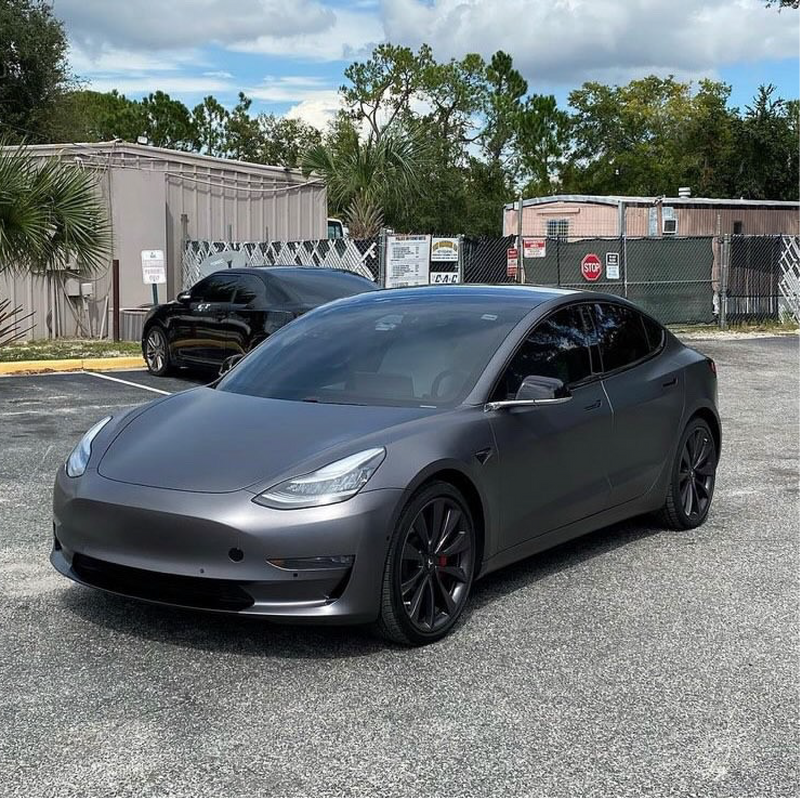 Gray Tesla car parked in a lot with trees and buildings in the background
