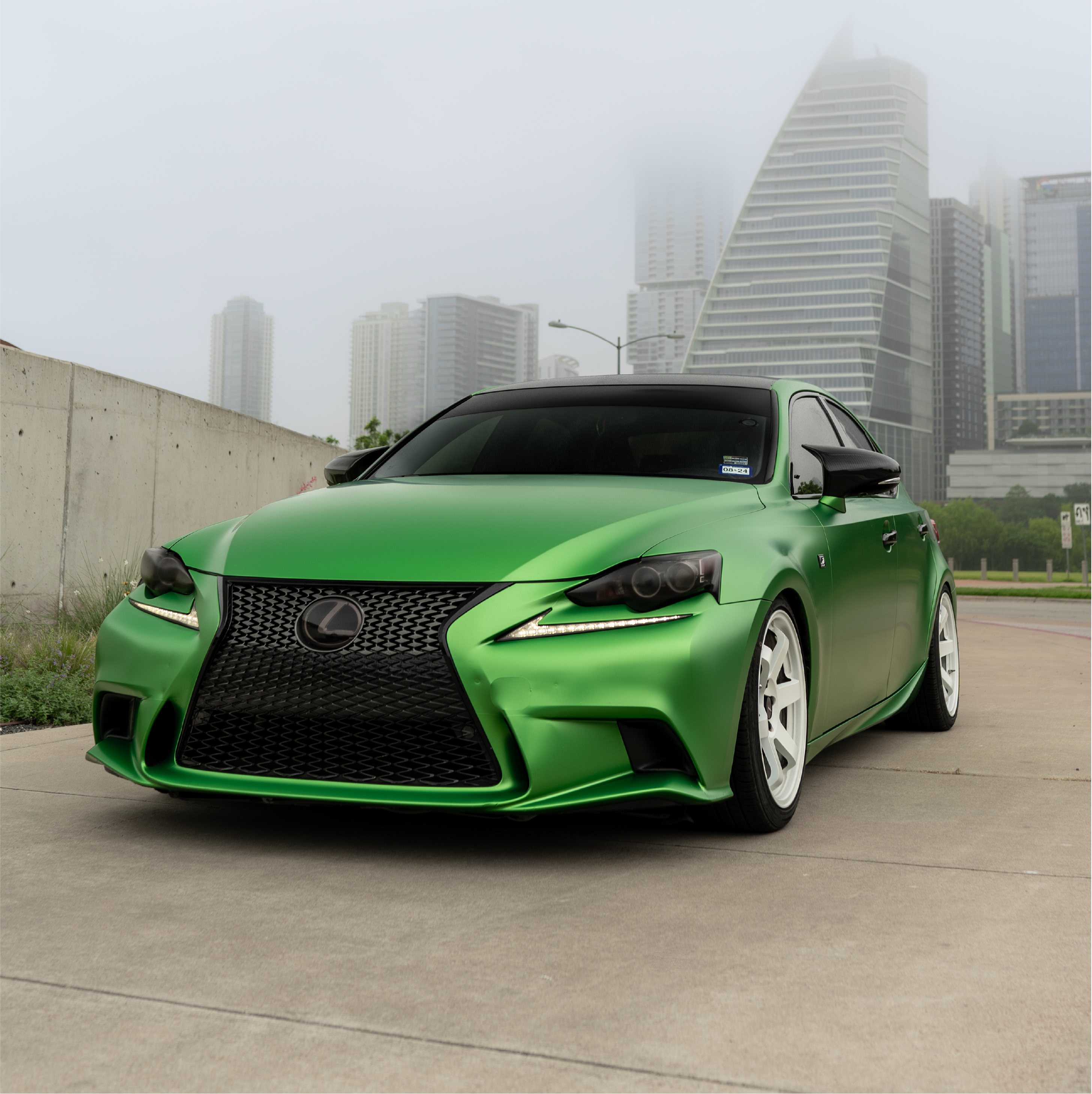 A modified Lexus IS sedan featuring a full Satin Cheetah Green Metallic vinyl car wrap and white wheels, set against a foggy city skyline.