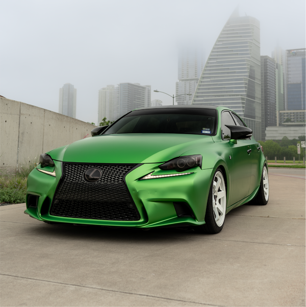 A modified Lexus IS sedan featuring a full Satin Cheetah Green Metallic vinyl car wrap and white wheels, set against a foggy city skyline.