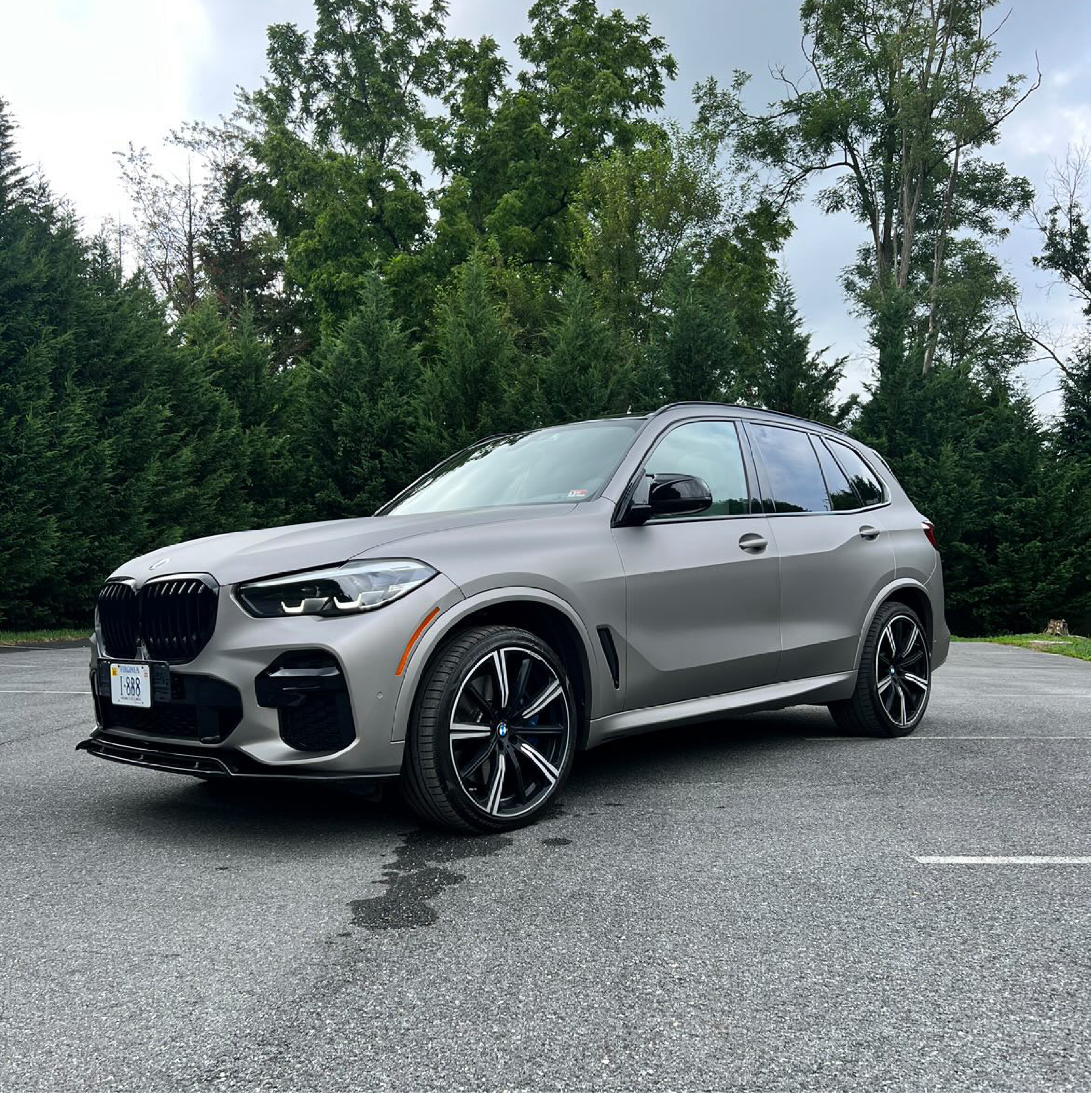 BMW X5 SUV wrapped in satin gray car wrap with black wheels, parked on asphalt in front of tall evergreen trees.