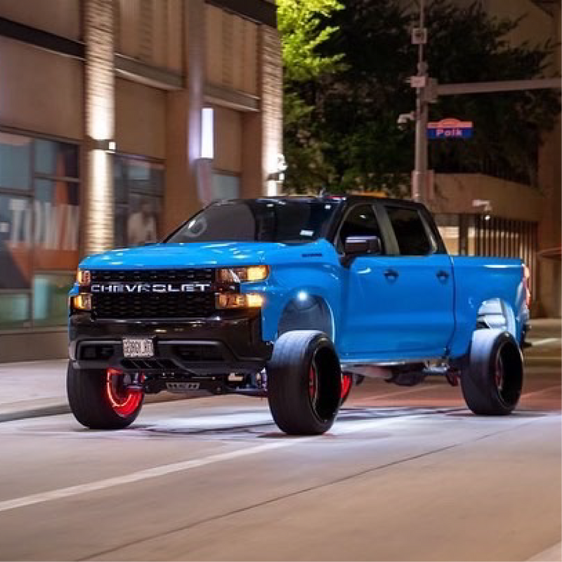 A truck wrapped in gloss light blue vinyl, driving on a road with buildings in the background.